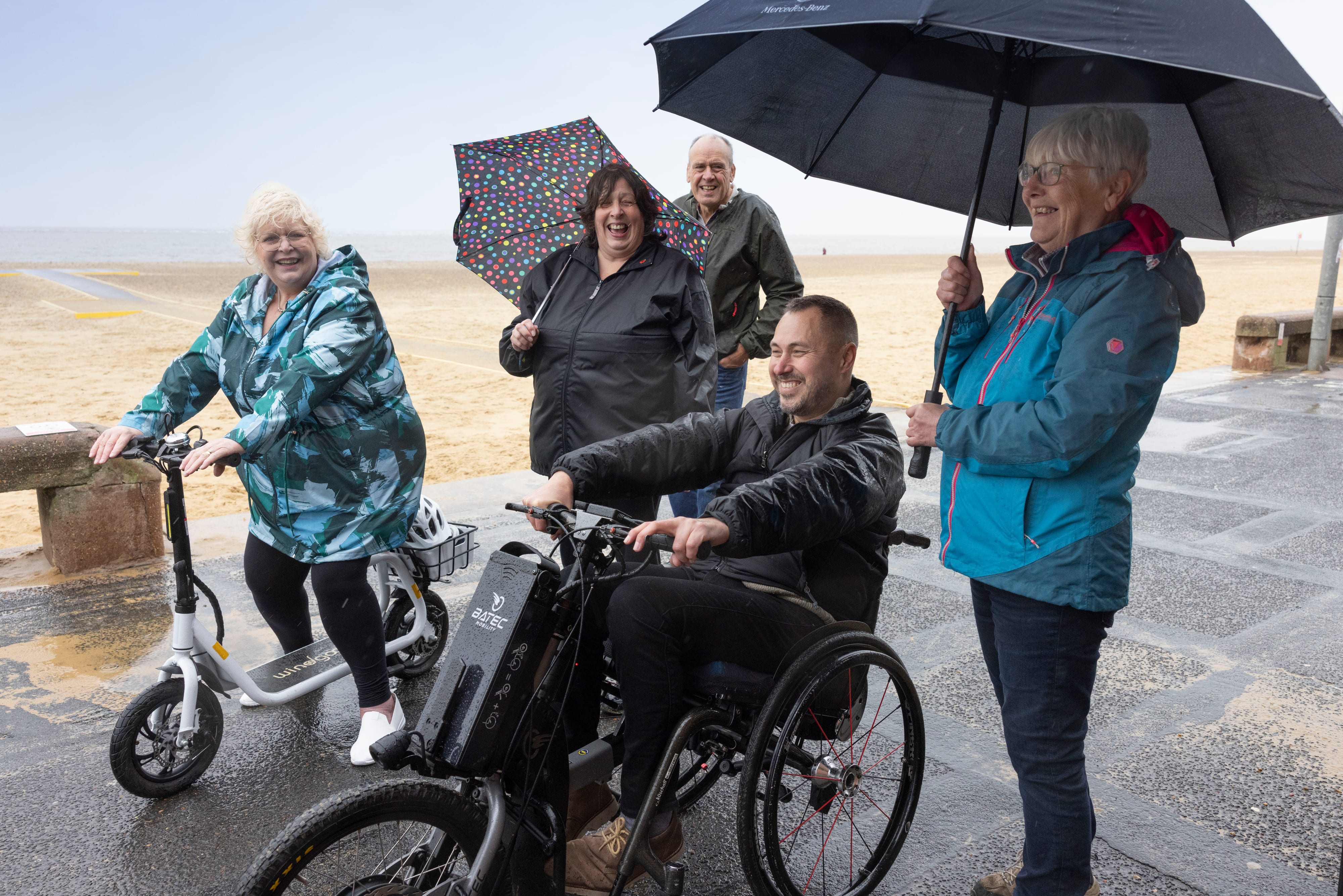 A man on a three wheeled wheelchair and a woman on a seated scooter on a board walk next to the beach smile. Three people stand behind.