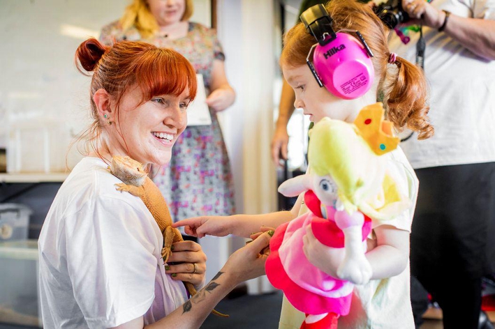 picture for projectStory: Child wearing ear phones holding doll touching a lizard held held by an adult. picture for projectStory: Child wearing ear phones holding doll touching a lizard held held by an adult.