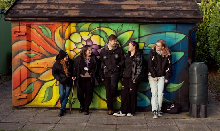 A group of young people standing outside in front colourful building community garden.