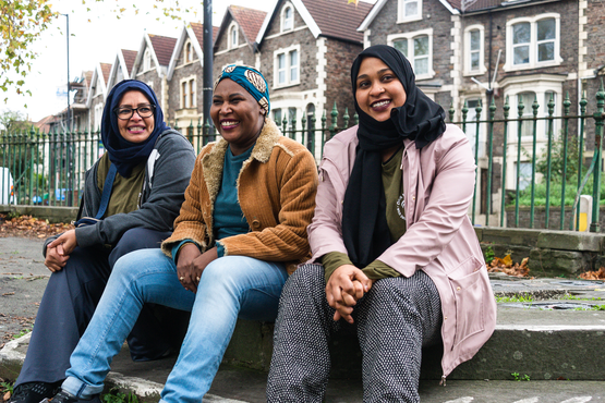 Three women  sit smiling on steps in front of a block of houses.