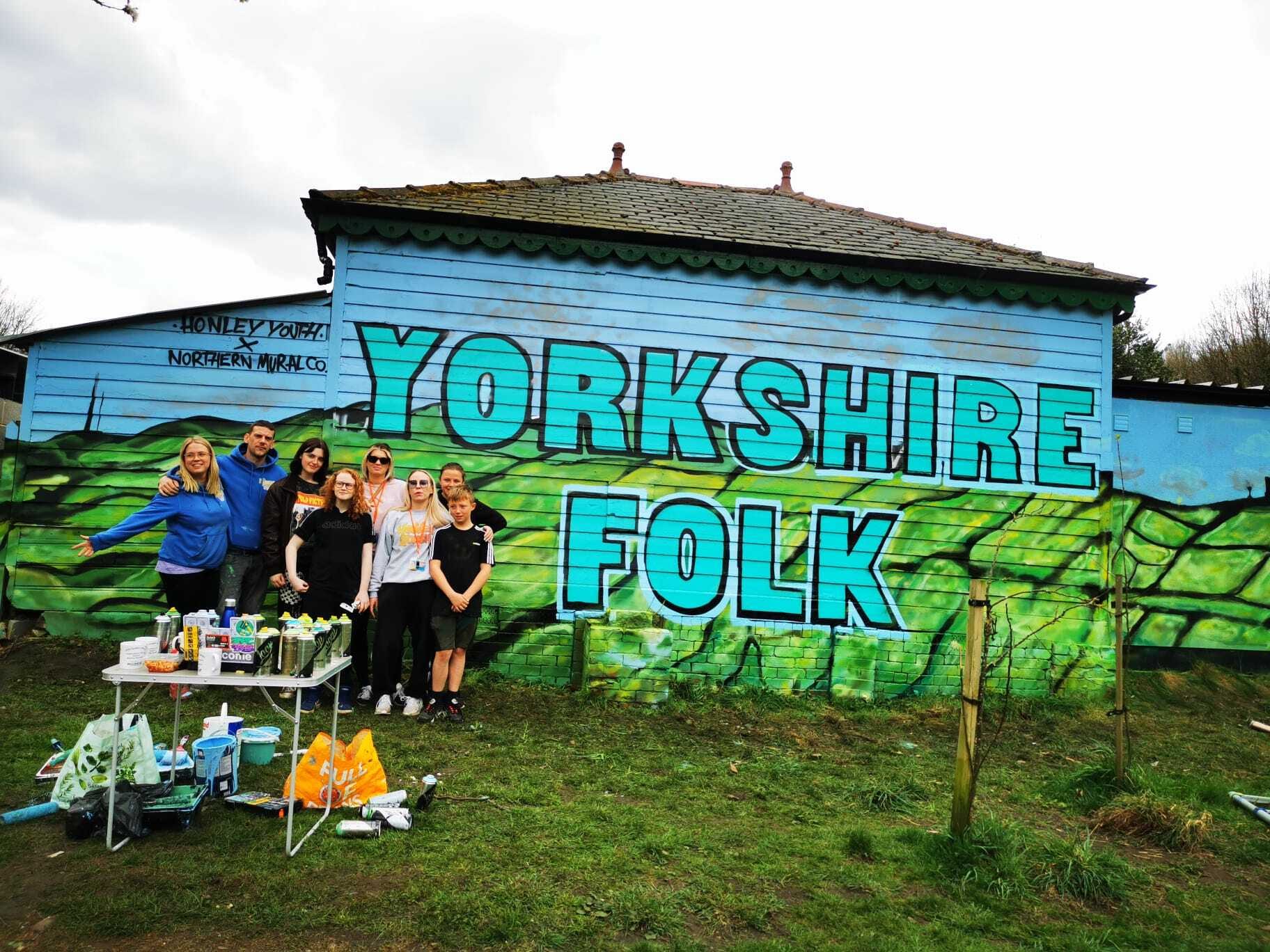 Young people stand in front of a building with graffiti reading Yorkshire Folk