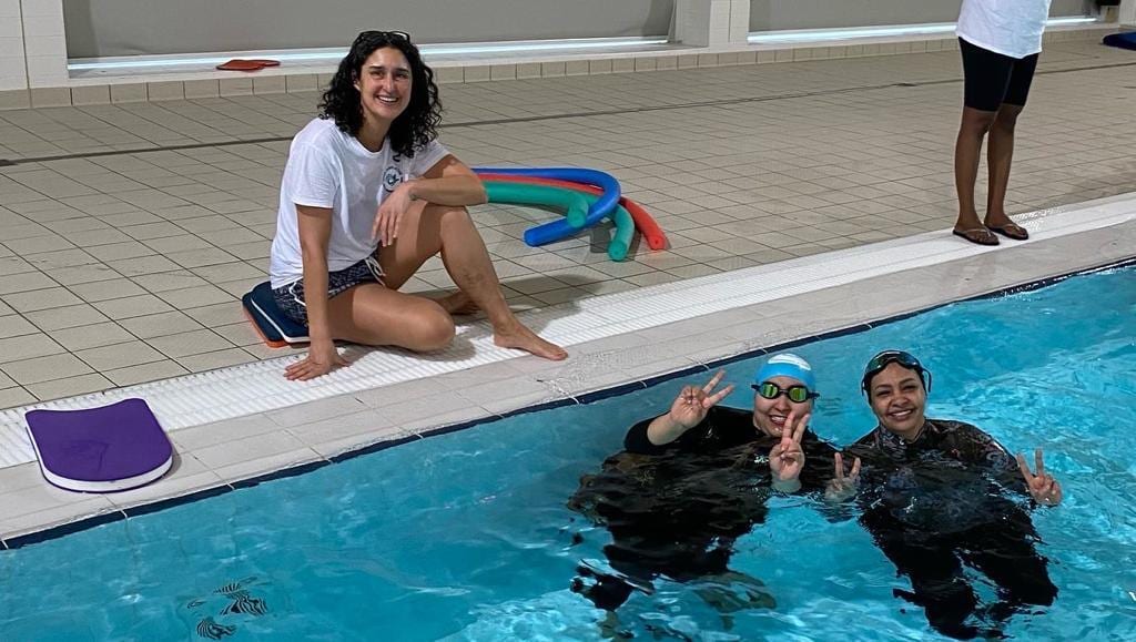 Two women in a swimming pool and a coach sat on the side